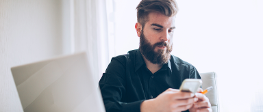 A person with short dark hair and a plaid shirt is sitting at a wooden table, looking at a silver laptop and holding a smartphone. They are taking a sip from a green cup of coffee. Another green cup is on the table behind their laptop. A window with a blurry yellow object outside is visible in the background.
