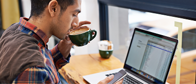 A man with a dark beard and a black shirt is sitting, looking down at a smartphone he's holding in both hands. A silver laptop is visible on a surface to his left, and a bright, light-colored background is behind him.