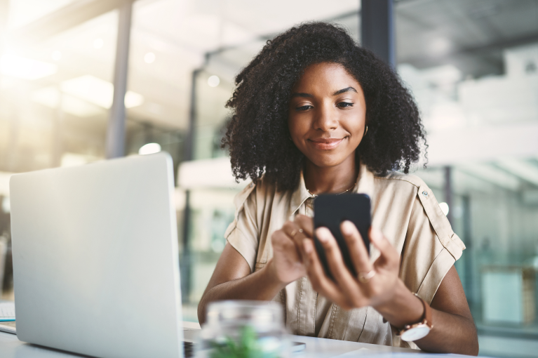 A woman looking at her phone, with a laptop in front of her.